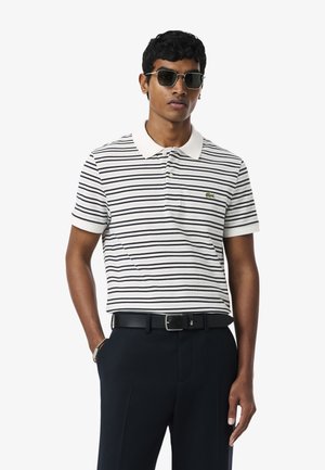 Giovane uomo con occhiali da sole neri, una polo bianca con strisce nere, pantaloni neri e cintura nera, in piedi con una mano in tasca.