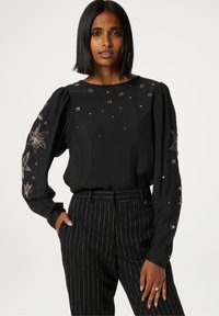 Zwarte blouse met pofmouwen en gedetailleerde geborduurde sterrenpatronen in zilver en goud. Gekoeld met zwarte broek met streepjes.