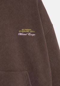 Sweat-shirt marron avec une poche avant et un texte jaune et rose portant la mention "No itinerary, no deadlines. Just a Mental Escape" sur la poitrine.
