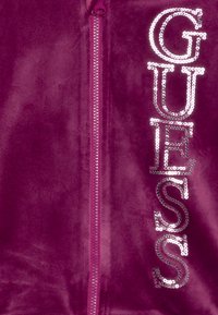 Giacca zip-up in velluto di un profondo viola con grandi lettere in paillettes argento "GUESS" lungo il lato sinistro. Il tessuto testurizzato aggiunge interesse visivo.