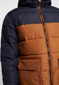 Kurtka pikowana z granatową górną częścią i brązową dolną; posiada kaptur, przedni zamek błyskawiczny oraz dwie boczne kieszenie.