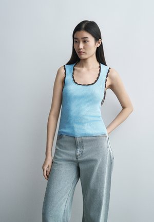 Młoda kobieta o długich czarnych włosach, ubrana w niebieski przylegający top bez rękawów z czarnymi wykończeniami oraz jasnoniebieskie dżinsy z wysokim stanem, stojąca z jedną ręką na biodrze.