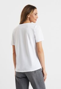 Witte katoenen t-shirt met korte mouwen, ronde halslijn en een subtiele logodetail op de achterkant. Gekleed met grijze denim broek.