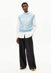 Lichtblauw gebreid vest met hoge kraag over een wit shirt, gecombineerd met zwarte wijde broek met krijtstrepen en zwarte schoenen met gele zolen.