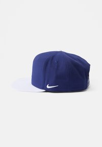 Gorra de béisbol azul y blanca con visera plana; presenta una punta blanca en contraste y el logotipo de Nike bordado en el lateral. Textura de tela de algodón.