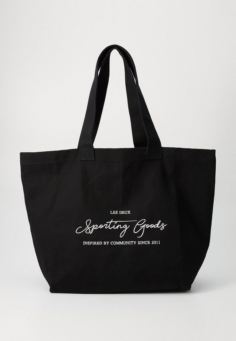 Svart kanvas tote-veske med doble robuste håndtak. Har hvit tekst: "LES DEUX Sporting Goods" og "Inspirert av fellesskapet siden 2011."