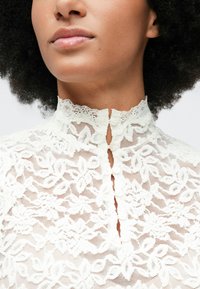 Nærbilde av en kvinne som har på seg en hvit bluse med høy hals i blonder med blomsterdesign og små frontknapper.