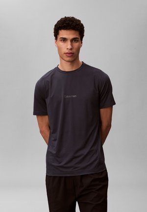 Ung mand med krøllet hår iført en tætsiddende mørkegrå Calvin Klein T-shirt, stående med hænderne bag ryggen mod en ensfarvet baggrund.