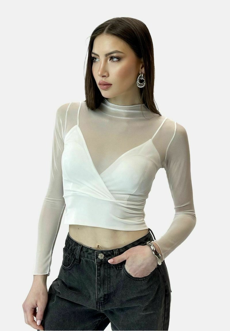 Hvid langærmet top med gennemsigtigt stofoverlæg, har en indpakningsdesign og tætsiddende bodice. Bæres med højtaljede sorte bukser.