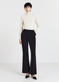 Schwarze, ausgestellte Hosen mit glatter Textur und hohem Bund, kombiniert mit einem cremefarbenen Rollkragenpullover. Das Modell trägt schwarze High-Heels.