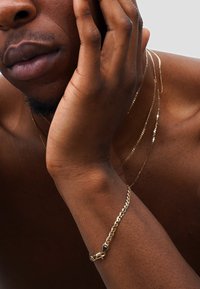 Gold chain necklace layered with additional thin gold chains, worn on bare skin. Visible bracelet with a unique clasp detail on wrist.