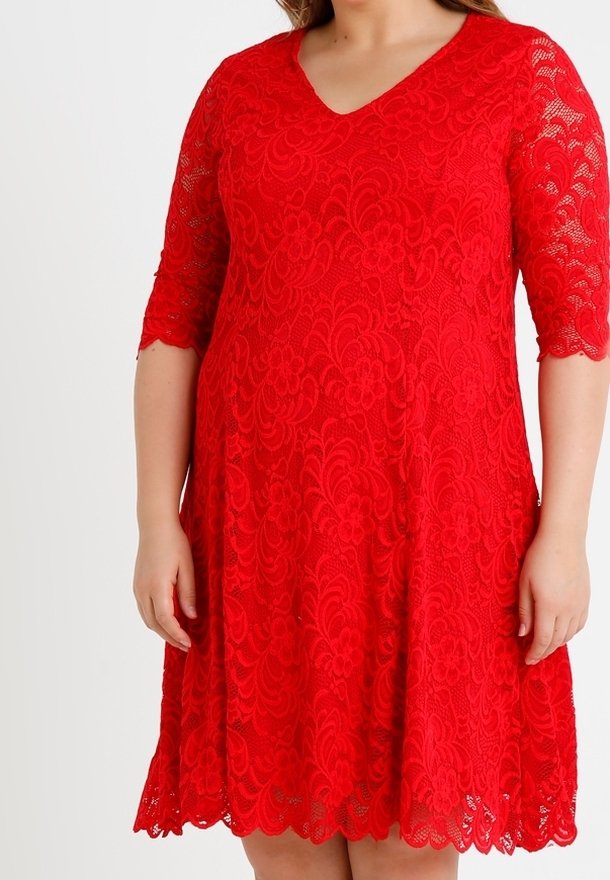 Femme portant une robe rouge en dentelle avec manches trois-quarts et un ourlet festonné, debout devant un fond blanc uni.