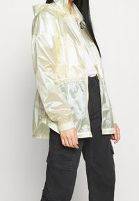 Ljusgul, blank vindjacka med huva, elastiska ärmslut och dragsko i midjan, bärs över en vit skjorta och svarta cargobyxor.