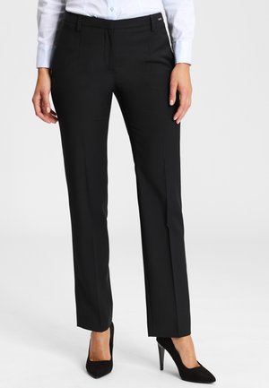 Femme portant un pantalon noir ajusté, une chemise blanche boutonnée et des chaussures à talons hauts noires, debout sur un fond clair uni.