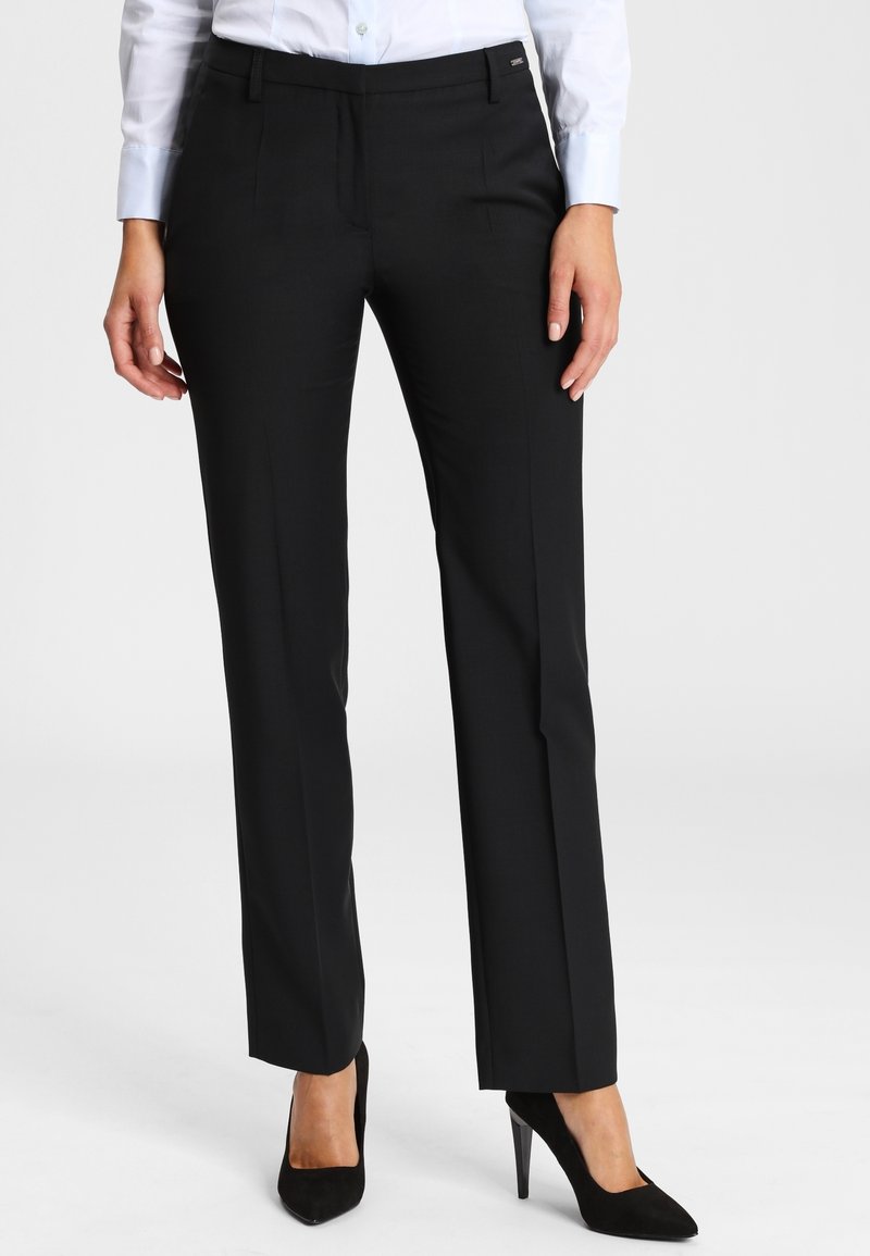 Femme portant un pantalon noir ajusté, une chemise blanche boutonnée et des chaussures à talons hauts noires, debout sur un fond clair uni.