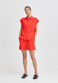 Camicetta rossa a maniche corte con bottoni e finitura testurizzata, abbinata a pantaloni corti rossi coordinati. L'outfit è completato da mocassini neri.