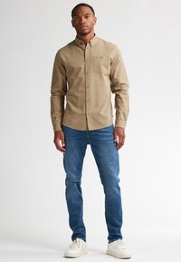 Lichtbruine knoopshirt met een borstzak, gecombineerd met blauwe jeans en witte sneakers. Eenvoudig ontwerp, standaard kraag en een gladde textuur.