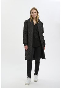 Manteau long matelassé noir avec capuche, doté d'une fermeture éclair et de poches latérales, associé à un pantalon noir et des baskets blanches.