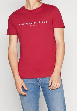Person trägt ein rotes Tommy Hilfiger T-Shirt mit Logo und dem Schriftzug "Est. 1985", kombiniert mit hellblauen Jeans vor einfachem Hintergrund.