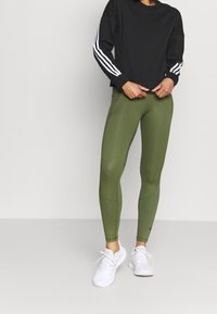 Felpa sportiva nera con strisce bianche sulle maniche, abbinata a leggings verde oliva. Le scarpe sono bianche con lacci e hanno un design sportivo.