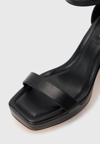 Zbliżenie na czarną skórzaną sandałkę z kwadratowym noskiem, pojedynczym paskiem z przodu i miękką wkładką na białym tle.