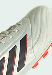 Scarpa da calcio bianca con parte superiore testurizzata, accenti a strisce nere e rosse e tacchetti in gomma arancione. Presenta chiusura con lacci e finitura liscia.