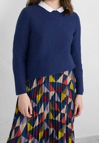 Maglione lavorato a maglia blu con colletto bianco, abbinato a una gonna plissettata multicolore con motivi geometrici in rosso, blu, giallo e crema.
