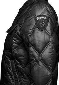 Giacca nera isolata con design a diamante trapuntato, con patch logo sulla spalla e tasche laterali, realizzata in un materiale lucido e liscio.