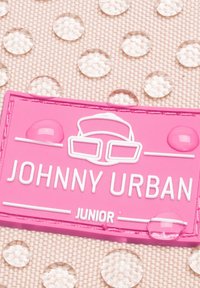 Rózsaszín gumicímke domborműben lévő "JOHNNY URBAN JUNIOR" felirattal és szemüveg grafikai dizájnnal, textúrált bézs háttéren, cseppekkel.