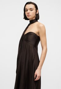 Γυναίκα με κοντά σκούρα μαλλιά που φοράει αμάνικο, πλισέ σκούρο καφέ φόρεμα με ταίριασμα στο λαιμό, στέκεται μπροστά σε απλό ανοιχτό φόντο.