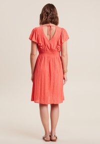 Korallenfarbenes Kleid mit kurzen, rüschierten Ärmeln, einer gerafften Taille und einem offenen Rücken. Der strukturierte Stoff weist vertikale Streifen mit subtilen metallischen Akzenten auf.