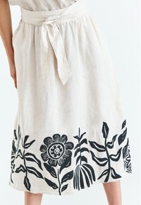 Linnedragt i lys beige med båndliv. Har sorte blomsterprint ved kanten, der viser forskellige blade og blomster.