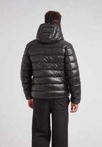 Giacca a vento nera imbottita con finitura lucida e cappuccio attaccato; presenta cuciture orizzontali e polsini elasticizzati. Pantaloni neri semplici.