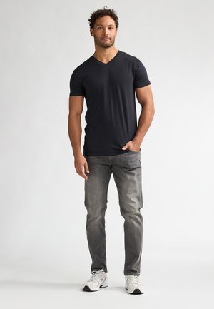 Zwart V-hals t-shirt van zachte stof, gecombineerd met lichtgrijze jeans. Model draagt witte sneakers en staat tegen een effen achtergrond.