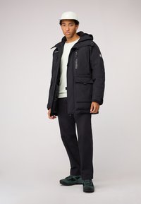Zwarte gewatteerde jas met capuchon, voorzien van meerdere ritszakken en een gestructureerde afwerking, gedragen over een lichtgroene trui en donkere broek.