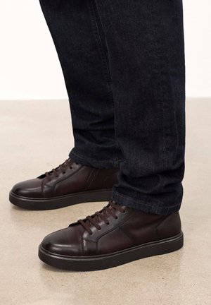 Donkerbruine leren hoge sneakers met platte veters, een gestructureerde zwarte rubberen zool en een minimalistisch ontwerp, gedragen met donkere jeans op een gladde ondergrond.