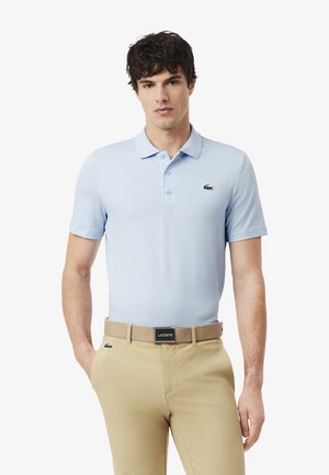 Uomo che indossa una polo azzurro chiaro e pantaloni beige con cintura abbinata, in piedi contro uno sfondo bianco, con le mani nelle tasche.