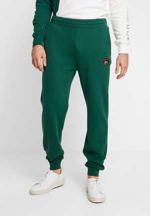 Homme portant un pantalon de jogging vert foncé avec des poignets élastiques et un petit patch Lacoste sur la cuisse gauche, assorti à des baskets blanches.