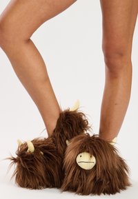 Des chaussons marron duveteux conçus pour ressembler à une créature, avec un visage souriant, des petites cornes et une texture douce. Revers autour de la cheville.