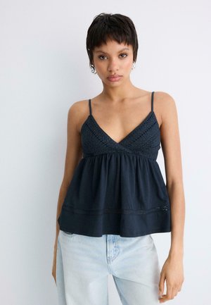 Młoda kobieta z krótkimi włosami, ubrana w ciemnoniebieski top na cienkich ramiączkach z haftem na szydełku oraz jasnoniebieskie dżinsy, stoi na tle gładkiej ściany.