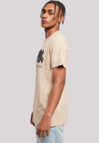 Beige cotton t-shirt with short sleeves, featuring a black graphic print on the front. Simple, relaxed fit. Light blue jeans visible below.