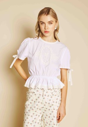 Fiatal nő szőke hajjal, fehér, fodros ujjú és peplum szegélyű blúzban, fehér virágos nadrággal, bézs háttér előtt.