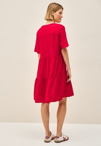 Rotes, knielanges Kleid aus leichtem Stoff mit kurzen Ärmeln und einem gestuften Rock. Tragen mit weißen Sneakers mit roten Akzenten.