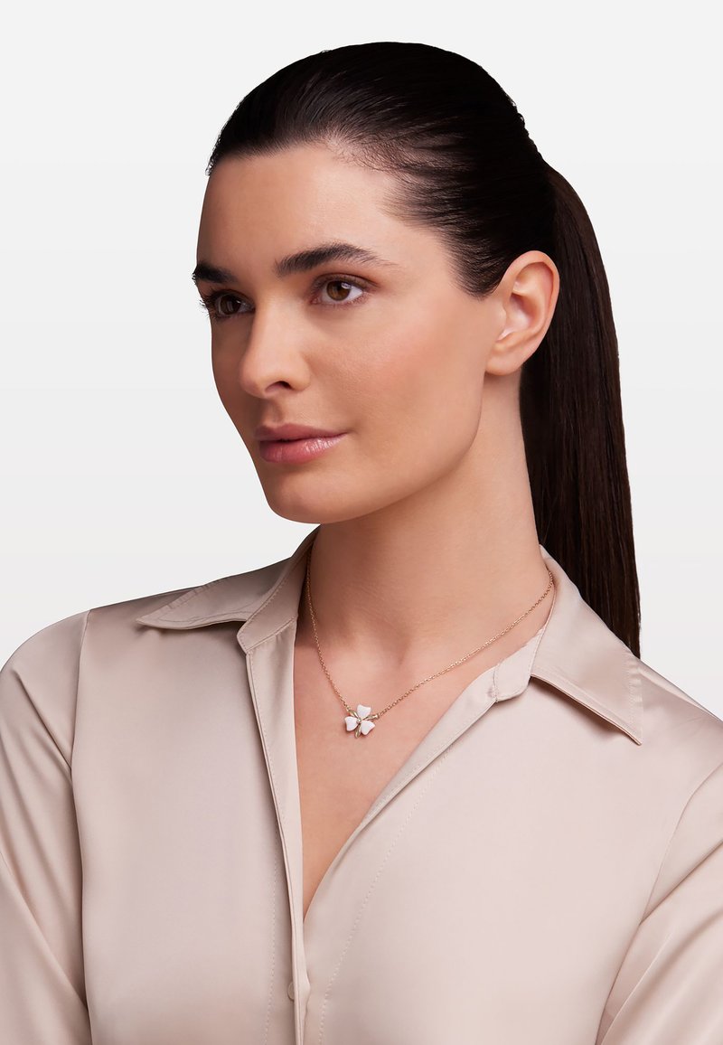 Collier en chaîne dorée avec un pendentif floral aux pétales blancs, porté par une personne en blouse claire sur un fond neutre.