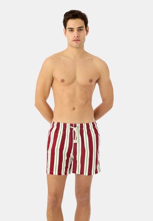 Junger Mann steht mit verschränkten Händen hinter dem Rücken und trägt rot-weiß-grün gestreifte Badeshorts vor einem einfarbigen Hintergrund.