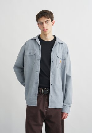 Νεαρός άνδρας με κοντά καστανά μαλλιά, φορώντας ένα ανοιχτό μπλε πουκάμισο πάνω από μαύρο μπλουζάκι και σκούρο καφέ παντελόνι με μαύρη ζώνη.