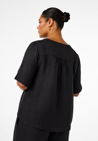 Black short-sleeve top with a relaxed fit, textured fabric, and a back ruffle detail. The neckline is round and hem is slightly curved.