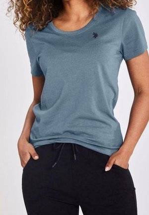 Personne aux cheveux bouclés portant une chemise à manches courtes bleu-gris ajustée et un pantalon noir avec cordon de serrage, les mains dans les poches, torse visible.