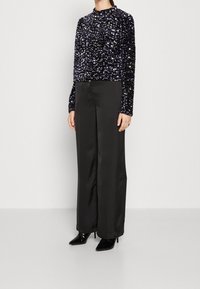 Top lungo a maniche di paillettes nere abbinato a pantaloni ampi in satin nero e tacchi a spillo neri, caratterizzato da un collo alto e un design aderente.