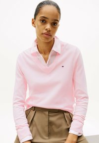 Helles rosa V-Ausschnitt-Pullover aus weichem Stoff, mit gerippten Bündchen und kleinem Logo. Dazu werden hellfarbige, plissierte Hosen getragen.